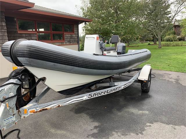 A gray inflatable boat with a white hull is mounted on a ShoreLand’r trailer in a driveway. The boat has seats and controls, and is parked near a building with a lawn and trees in the background.