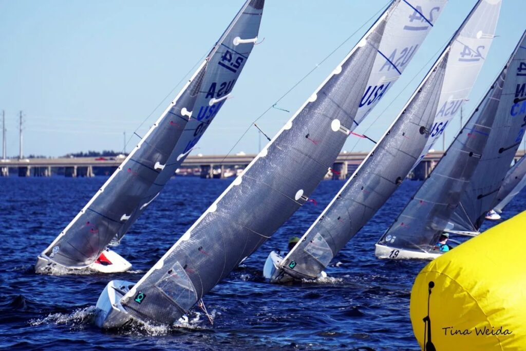 Four sailboats with silver sails racing closely on blue water near a bright yellow buoy, with a bridge and clear sky in the background. The image is signed 'Tina Weida' in the corner.