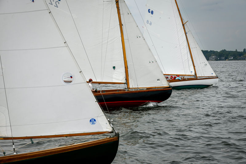 Three sailboats with white sails race side by side on a cloudy day, their bows cutting through the rippled water, with a distant shoreline visible in the background.