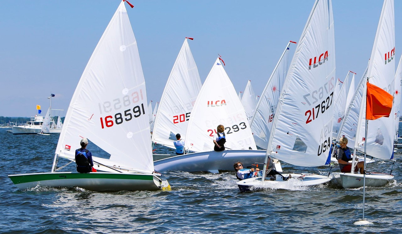 Several small sailboats with white sails compete in a regatta on a sunny day. Sailors, mostly youth, navigate around an orange marker buoy, with choppy water and other boats visible in the background.