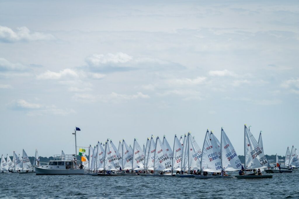 A large group of small sailboats with white sails lines up on the water near a boat displaying flags, under a partly cloudy sky.