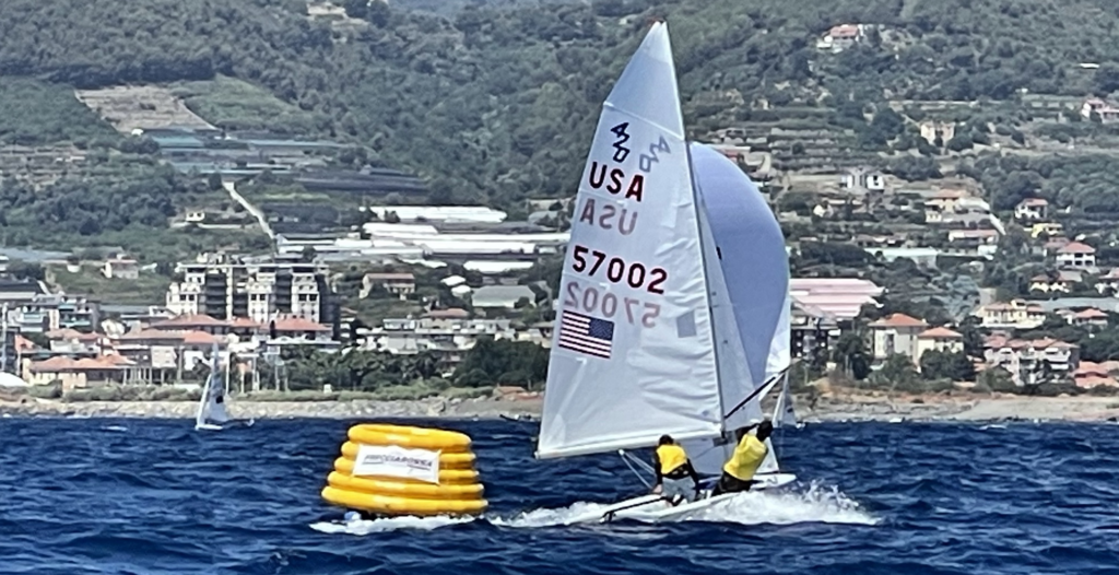 Two sailors in yellow gear race a sailboat marked 'USA 57002' around a yellow inflatable buoy on blue water, with a coastal town and green hills visible in the background.
