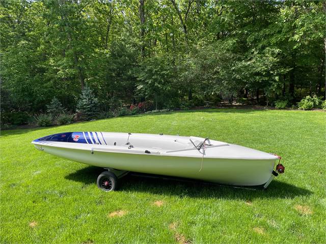 A small white sailboat with blue and red markings rests on a grassy lawn, supported by a small two-wheeled dolly, with green trees in the background.