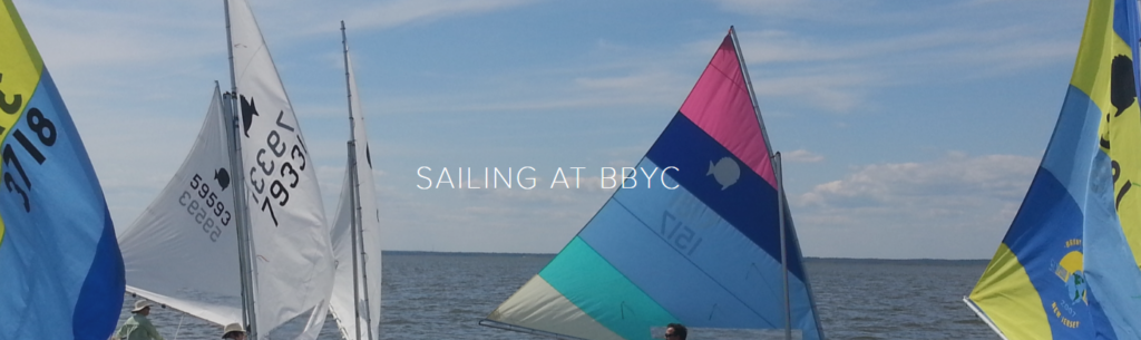 Colorful sailboats with unique sail patterns and numbers are gathered on the water under a blue sky. The text 'SAILING AT BBYC' is centered over the image.