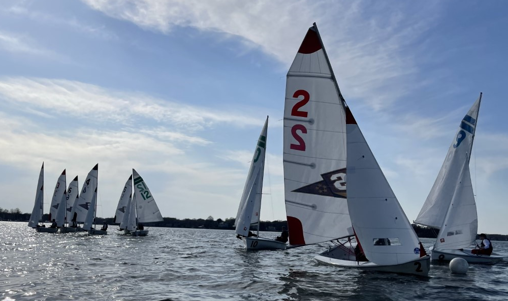 Several small sailboats with numbered sails race on a lake under a partly cloudy sky. The sun reflects on the water, and a tree-lined shore is visible in the background.