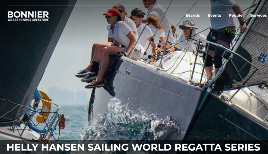 A group of sailors sit on the edge of a sailboat, leaning out over the water during a regatta. The boat is tilted, and another sailboat is visible nearby. Text overlays promote the Helly Hansen Sailing World Regatta Series.