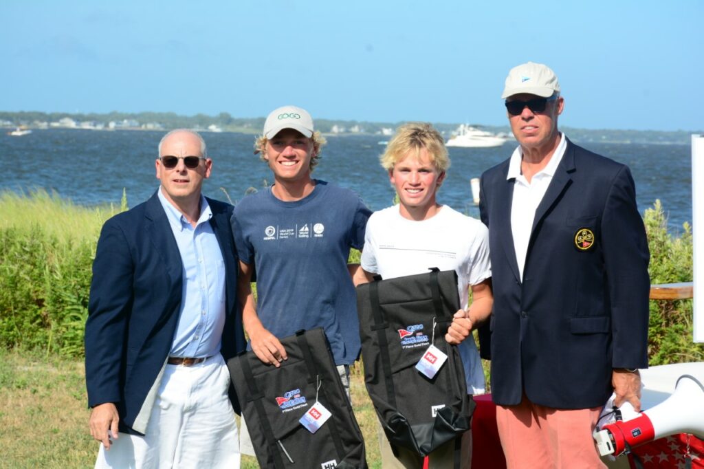 Four men stand outdoors near a body of water, two in blazers and sunglasses, two younger men in casual clothes holding black sailing bags, all smiling for a group photo on a sunny day.
