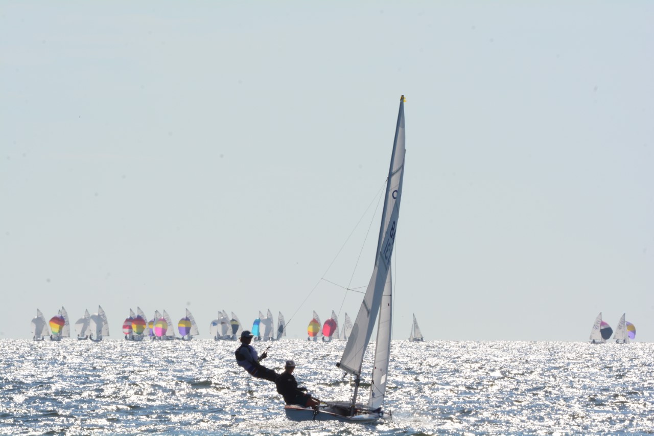 Two people sail a small boat on a sparkling, sunlit sea, leaning out to balance. In the distance, many sailboats with colorful sails are visible against the bright sky and water.