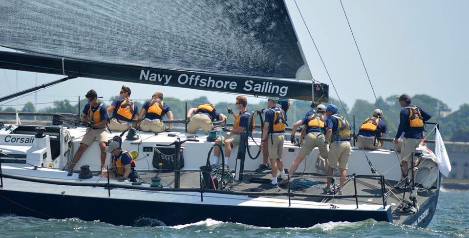 A team of sailors in matching uniforms works together on a sailboat labeled 'Navy Offshore Sailing' as it travels across the water on a sunny day.
