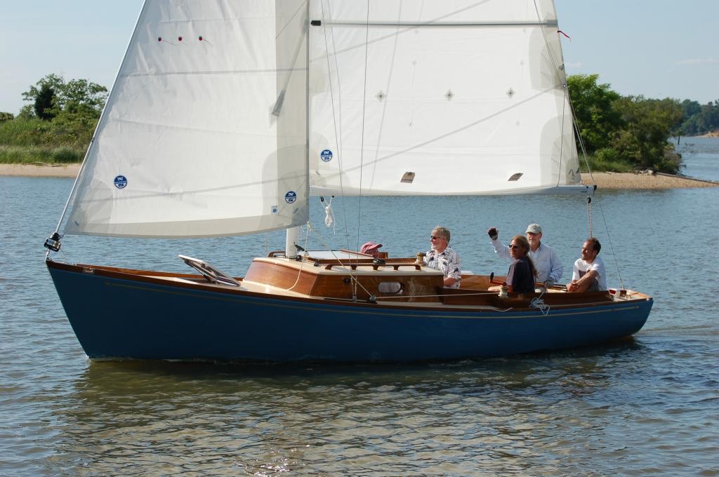 Four people are sailing a small blue and wooden sailboat on calm water near a sandy shoreline with trees and greenery in the background under a clear sky.