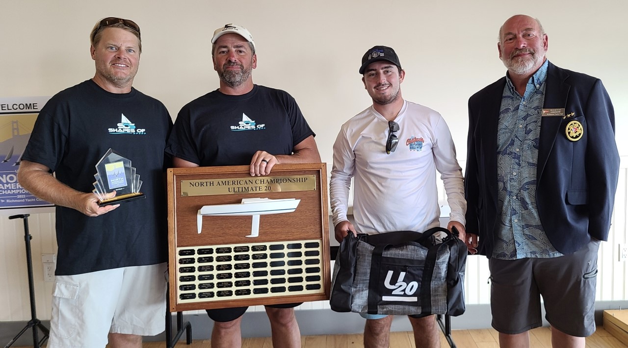 Four men stand indoors posing for a photo; two are holding trophies, one holds a large wooden plaque labeled "North American Championship Ultimate 20," and the fourth stands at the end wearing a blazer.