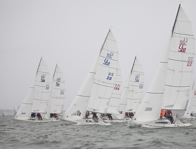 Several white sailboats with “420” on their sails race closely together on a grey, choppy body of water under an overcast sky. Sailors wearing life jackets are visible on each boat.