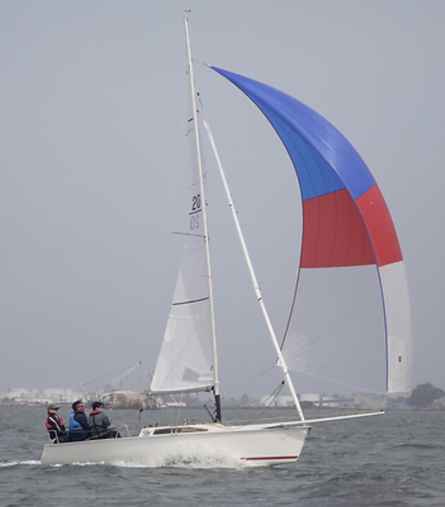 A small sailboat with three people onboard glides over the water. The boat has a blue, red, and white spinnaker sail raised, with a foggy shoreline visible in the background.
