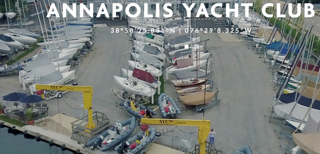 Aerial view of the Annapolis Yacht Club boatyard, showing sailboats lined up on land, two yellow boat hoists labeled 'AYC,' and docked boats near the water. Text at the top reads “ANNAPOLIS YACHT CLUB” with coordinates below.