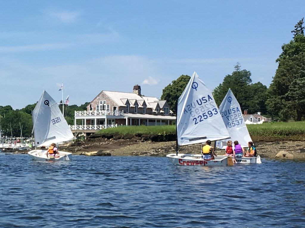 Children sailing small boats near a grassy shoreline with a white, multi-story clubhouse and trees in the background on a sunny day. The boats' sails display numbers and the letters 'USA.