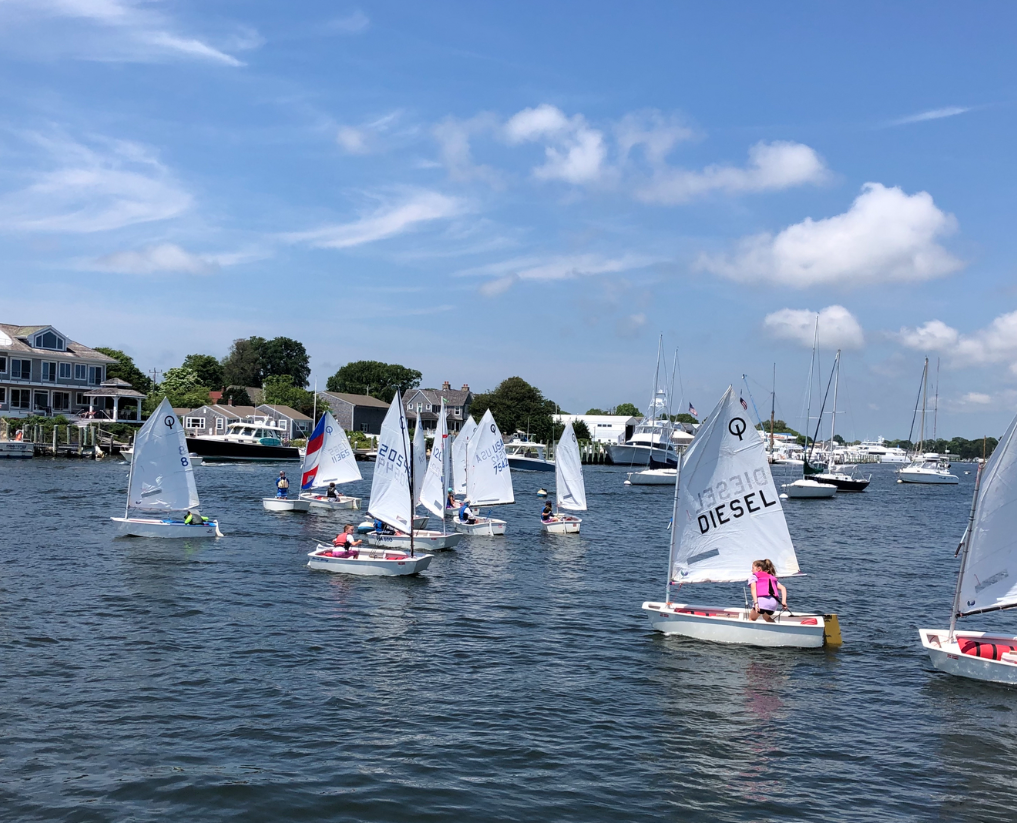 Small sailboats with children sailing on a calm harbor, with houses, trees, and moored boats visible in the background under a blue sky with scattered clouds.