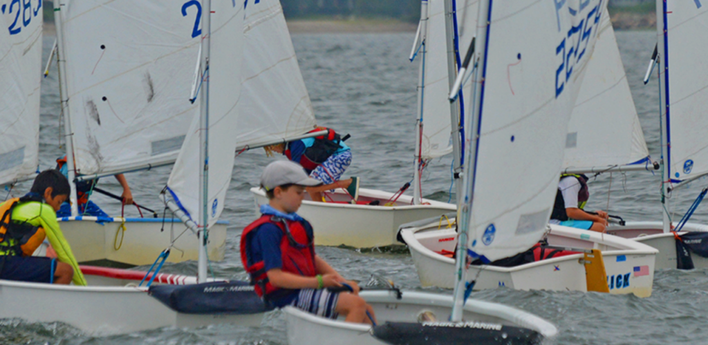 Children wearing life vests and caps are sailing small white dinghies close together on a body of water, with a blurred shoreline visible in the background.