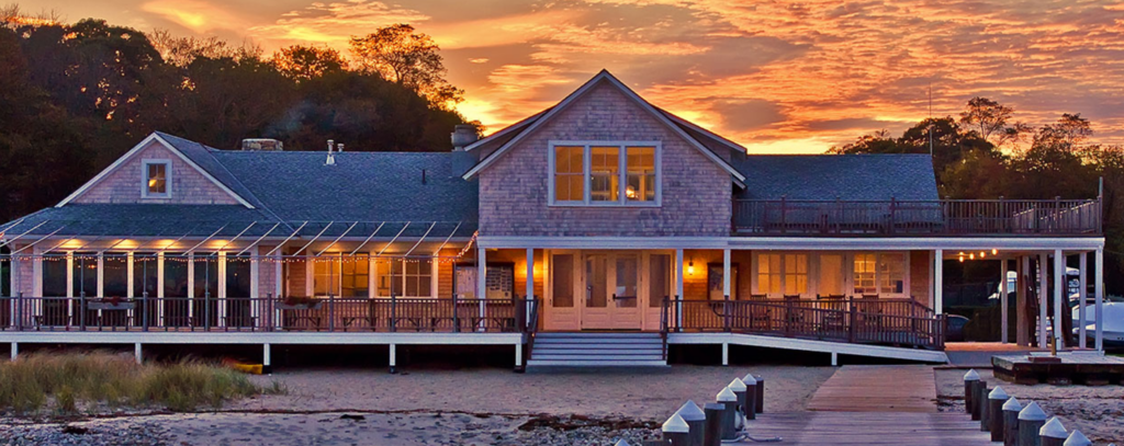 A large wooden beach house with a wraparound porch, lit warmly from inside at sunset. A boardwalk leads up to the house, and trees are silhouetted against a colorful sky in the background.