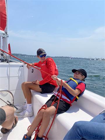 Two people in red shirts and life jackets are sailing on a boat. One is pulling a red rope while the other sits nearby, both wearing sunglasses and caps. The water and distant shoreline are visible in the background.