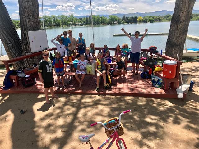 A group of children and adults pose and smile on a wooden deck by a lake, surrounded by trees. There are coolers, bags, and a child's bicycle in the foreground, with mountains and blue sky in the background.