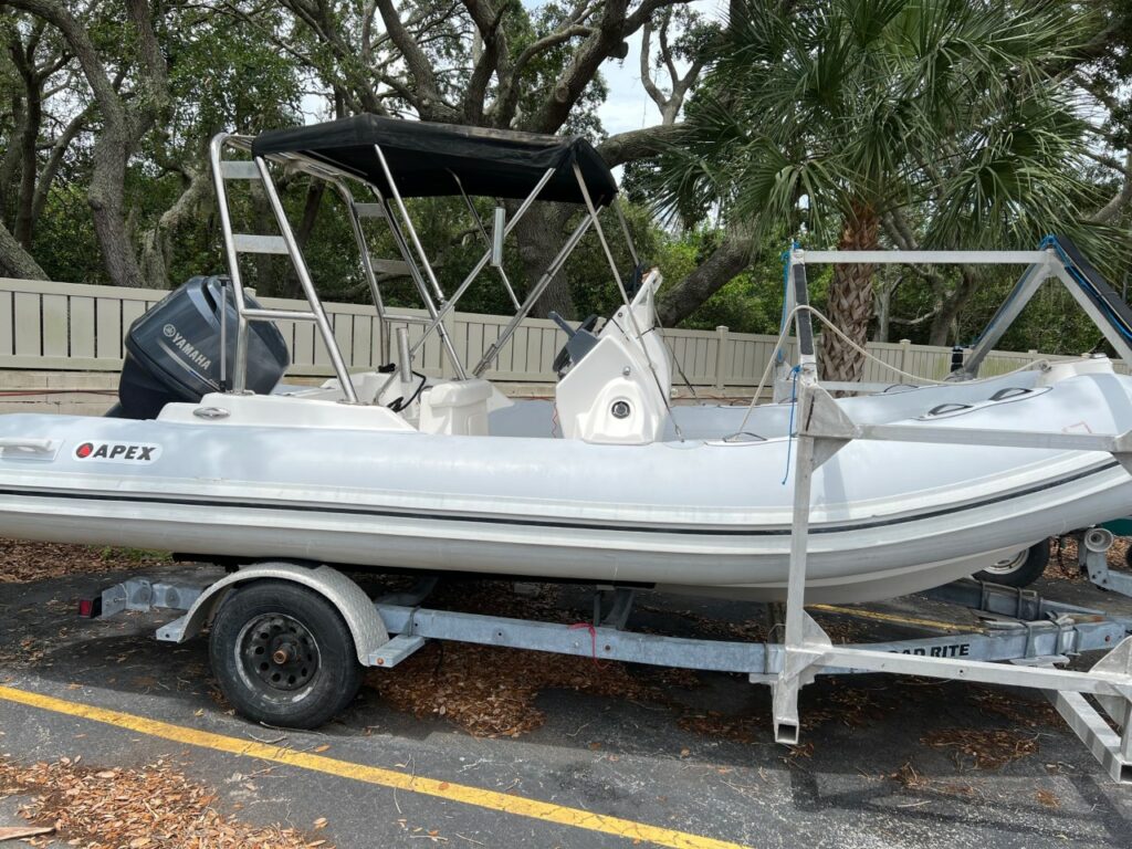A white inflatable Apex boat with a Yamaha outboard motor is on a metal trailer in a parking lot. The boat has a black canopy and is surrounded by trees and a light-colored fence in the background.