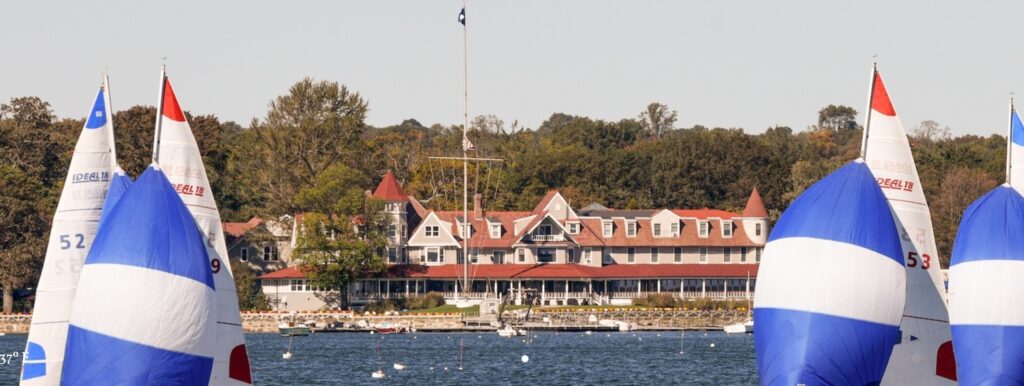 Several sailboats with blue and white sails race on a lake in front of a large, historic-looking building with a red roof, surrounded by trees and a clear sky.
