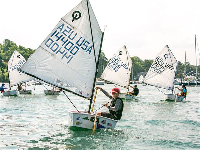 Several young sailors race small sailboats on a calm body of water, each boat displaying sail numbers. Trees and additional boats are visible in the background on a sunny day.