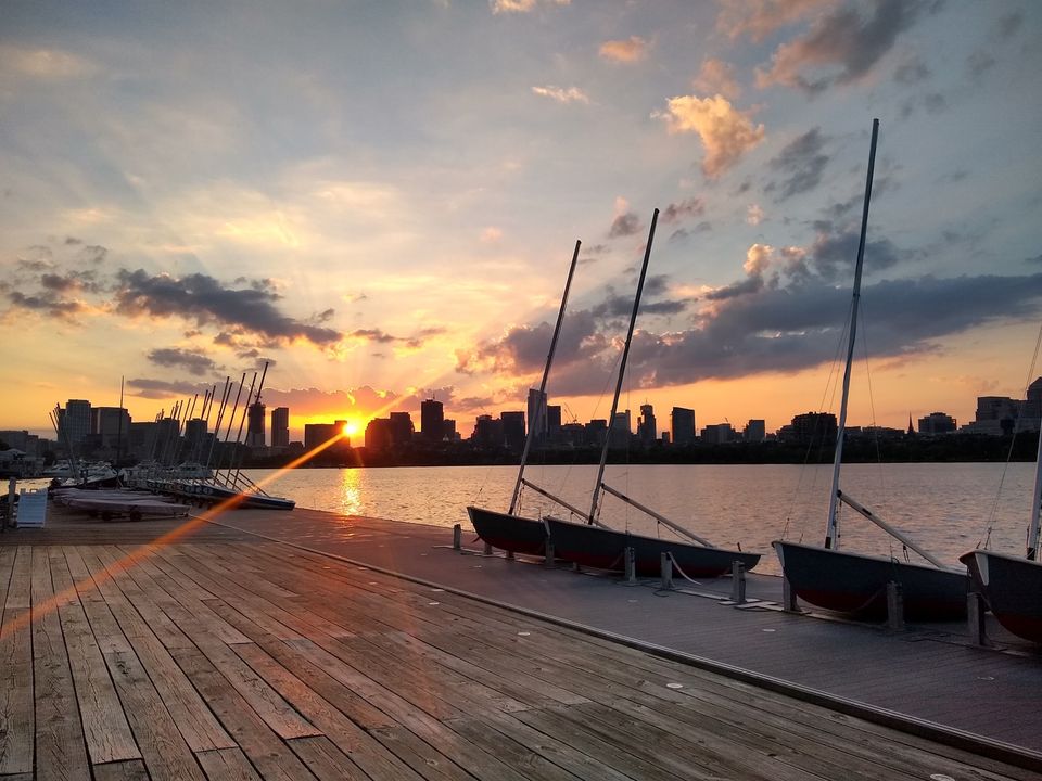 Sailboats docked on a wooden pier at sunset, with a city skyline in the background and colorful clouds in the sky reflecting on the water.