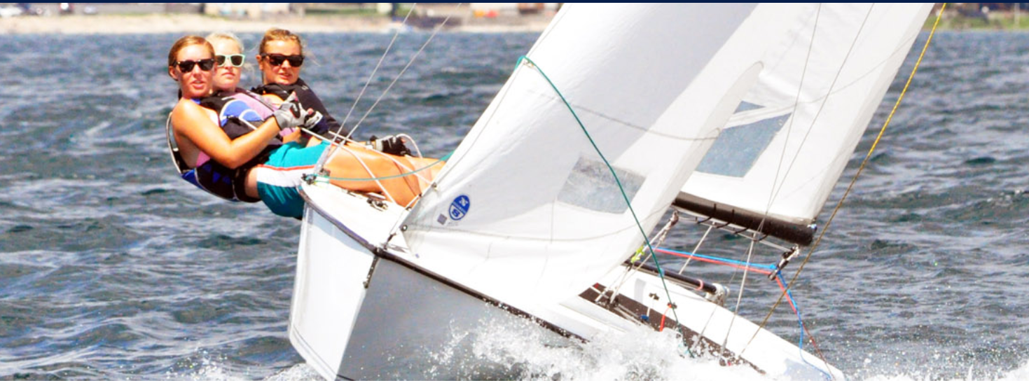 Three people wearing sunglasses and life jackets lean out from the side of a small white sailboat as it tilts on the water, with the sails catching the wind and the shoreline visible in the background.