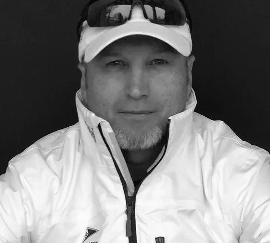 A man wearing a white jacket, white baseball cap, and sunglasses resting on his head, looking at the camera with a neutral expression against a dark background.
