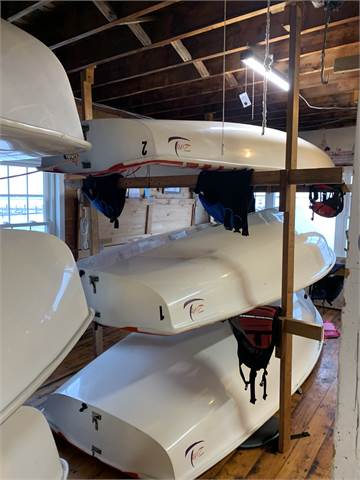 Three white kayaks are stacked on wooden racks in a wooden-floored boathouse. Life jackets hang on the racks, and sunlight streams in through a window on the left. The ceiling has exposed beams and lighting.