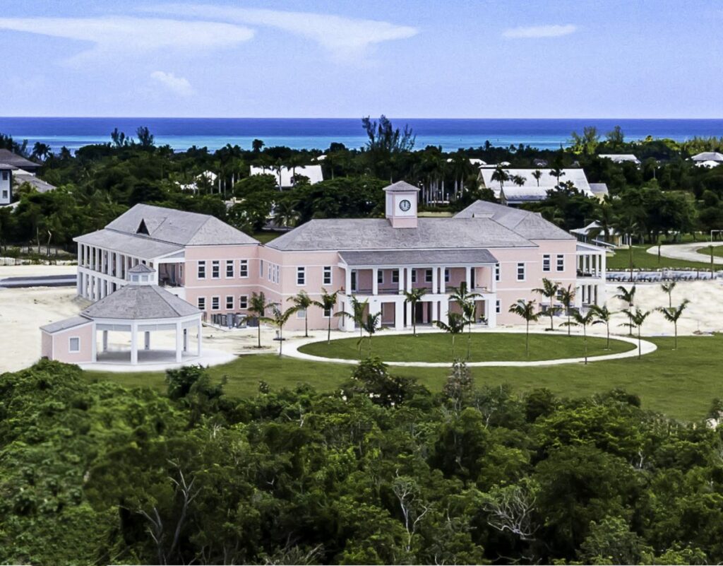 Aerial view of a large, pink building with a clock tower, surrounded by palm trees and greenery, with the ocean and blue sky in the background.