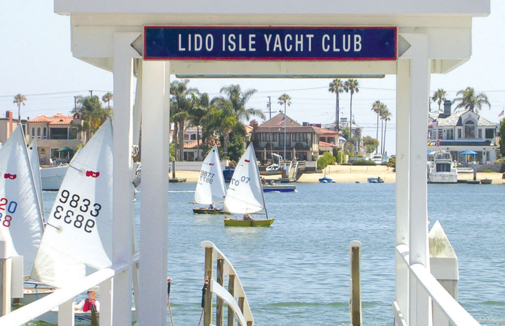 A dock entrance with a sign reading “Lido Isle Yacht Club” overlooks a bay with small sailboats and waterfront houses lined with palm trees in the background.