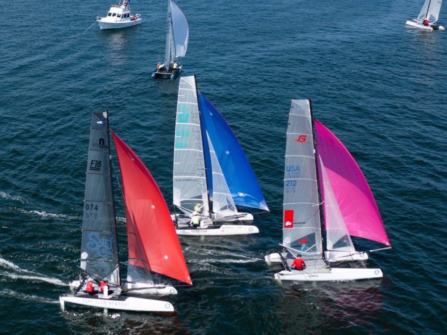 Three sailboats with bright red, blue, and pink sails race closely together on open water, with two other boats visible in the background. The water is deep blue and the scene is lively.