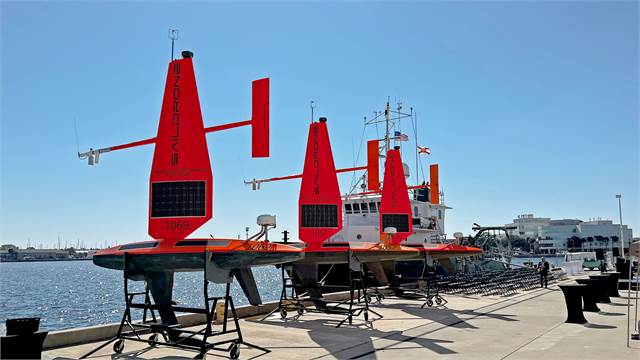 Three bright orange autonomous ocean drones with solar panels are lined up on dockside stands near a harbor, with a large ship and city buildings in the background under a clear blue sky.