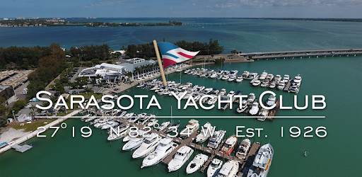 Aerial view of Sarasota Yacht Club marina with many docked boats, a large flag, and surrounding water. Text reads: “Sarasota Yacht Club 27°19’N 82°34’W Est. 1926.”.