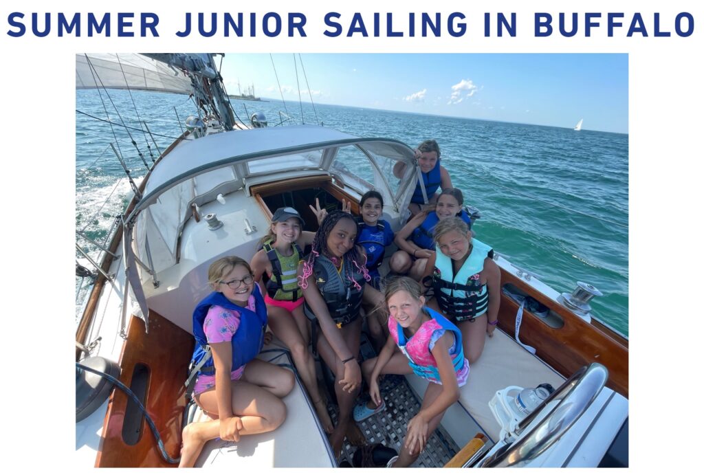 A group of smiling kids wearing life jackets sit together on a sailboat under sunny skies with 'SUMMER JUNIOR SAILING IN BUFFALO' written above them. The boat is on open water with a sail and blue sky in the background.