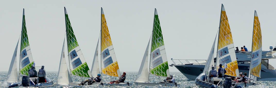 Several small sailboats with colorful, geometric-patterned sails—yellow and green—float on calm water, with people onboard. A larger motorboat with more people is visible in the background. The sky is clear.