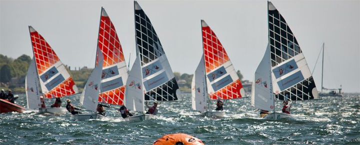 Several small sailboats with colorful sails are racing on a choppy body of water, with sailors visible in each boat and a large orange buoy in the foreground. Trees and buildings are visible in the background.