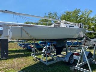 A white sailboat is secured on a trailer in a grassy outdoor area, surrounded by other boats and trees under a clear blue sky.