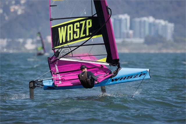 A person sails a hydrofoil WASZP sailboat with a purple and blue sail, gliding above the water with city buildings and hills visible in the background.