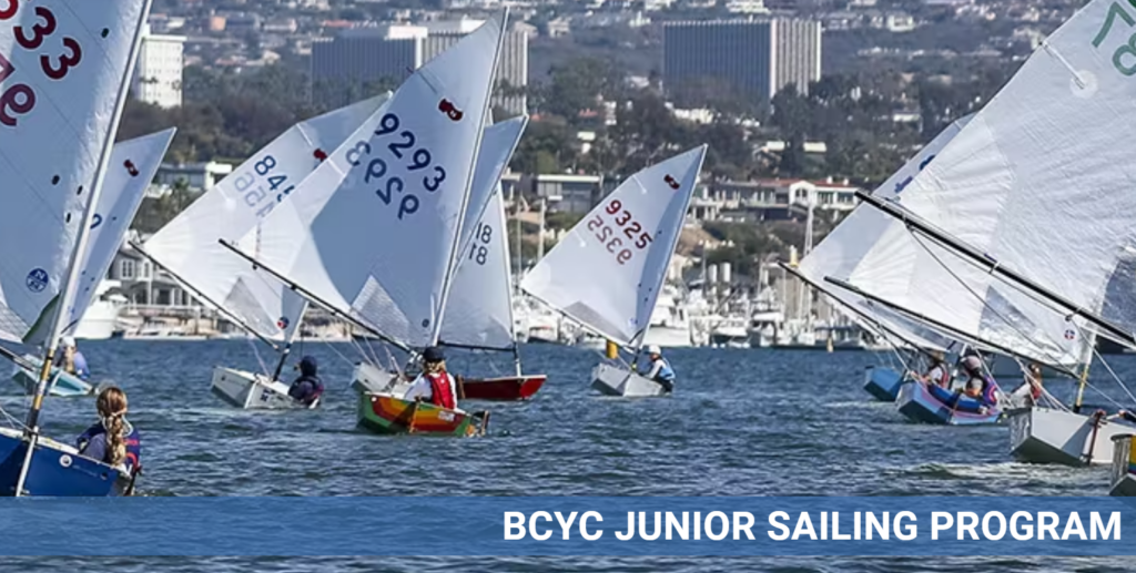 Children sail small boats on a sunny day in a marina, with city buildings and hills in the background. Text at the bottom reads 'BCYC JUNIOR SAILING PROGRAM.