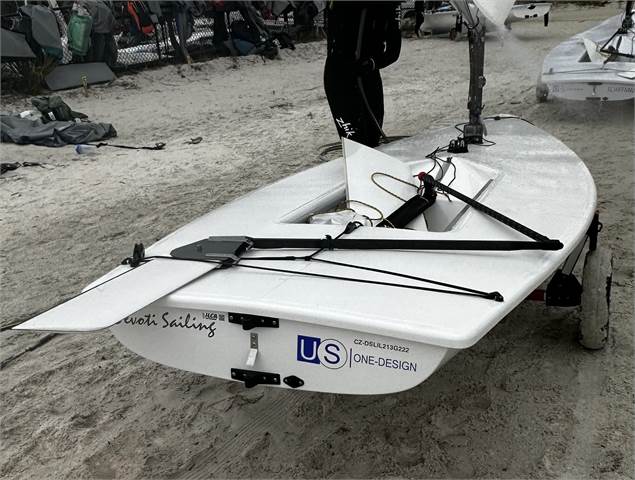 A small white sailboat on a sandy beach, equipped with a rudder, tiller, and ropes. The boat has "US One-Design" and "Swi Sailing" labels on its stern, and other boats and equipment are visible in the background.