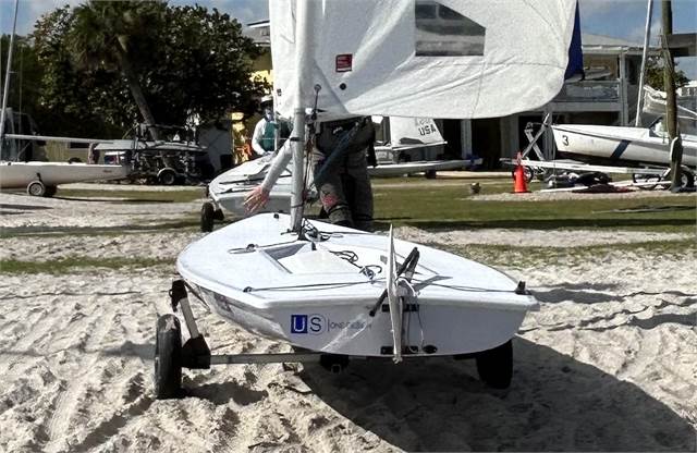 A small white sailboat on a dolly is on sandy ground near grass, with other boats and trees in the background. A person partially visible behind the sail is moving the boat.