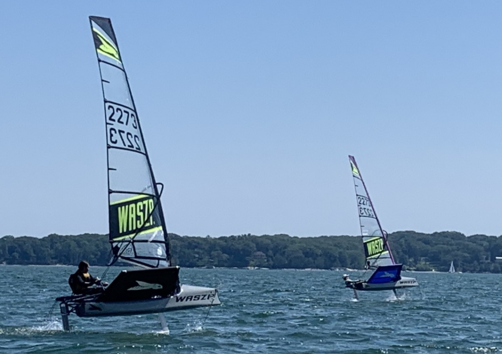 Two people sail on small hydrofoil sailboats labeled 'WASZP' across a lake, with trees and a clear blue sky in the background. The boats are elevated above the water on their foils.
