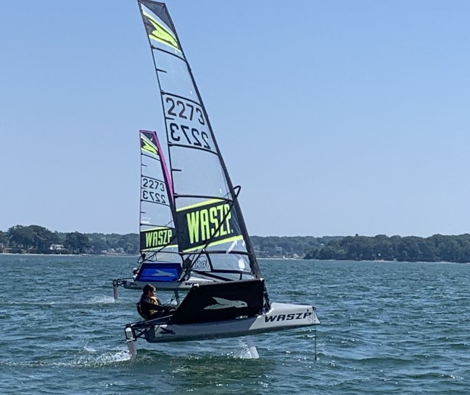 Two small sailboats with "WASZP" sails glide over the water on a sunny day, with one boat in the foreground and another behind it. A shoreline with trees is visible in the background.