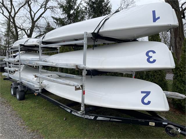 A trailer parked on grass carries five stacked white sailboats, each labeled with a blue number. Trees and shrubbery are visible in the background.