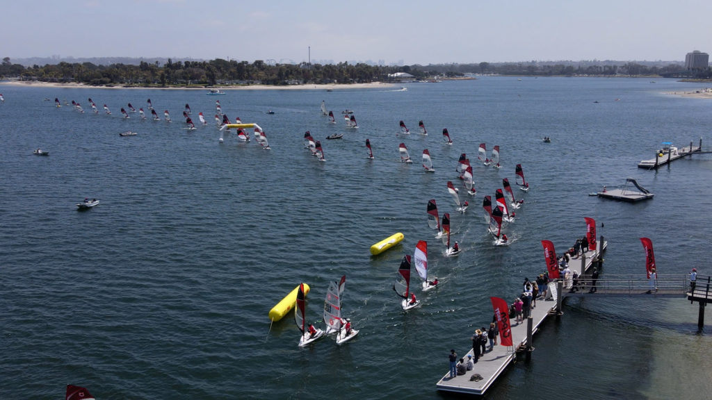 A group of windsurfers with red and white sails race on a blue lake, passing yellow buoys near a dock with onlookers and red flags. A tree-lined shore and buildings are visible in the background.