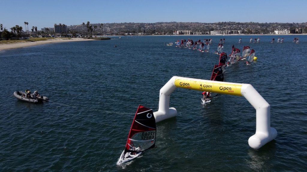 Small sailboats race through an inflatable arch on a sunny bay, with a motorboat nearby and many more sailboats visible in the distance. Apartment buildings and palm trees line the shore under a clear blue sky.