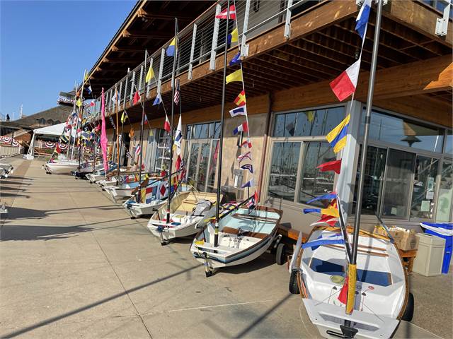 Small sailboats decorated with colorful nautical flags are lined up on a concrete dock beside a wooden building on a sunny day.